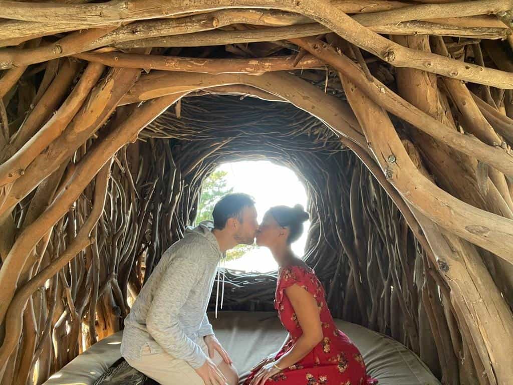 Couple kissing in a magical woven branch tunnel suite at a romantic hotel in the USA. The intimate wooden cocoon features a comfortable bed, natural lighting, and creates a perfect sanctuary for couples seeking unique luxury accommodations. This nature-inspired hideaway exemplifies the enchanting experiences available at top romantic hotels across the United States.