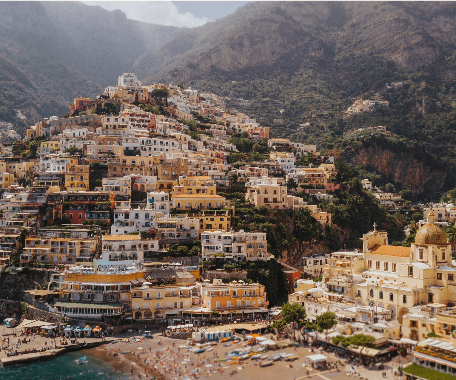 Stunning panoramic view of Positano on the Amalfi Coast, Italy, showcasing colorful pastel buildings cascading down steep cliffs to the beach below. A church with a golden dome is visible among the Mediterranean architecture, with dramatic mountains forming a backdrop against the turquoise waters of the Tyrrhenian Sea.