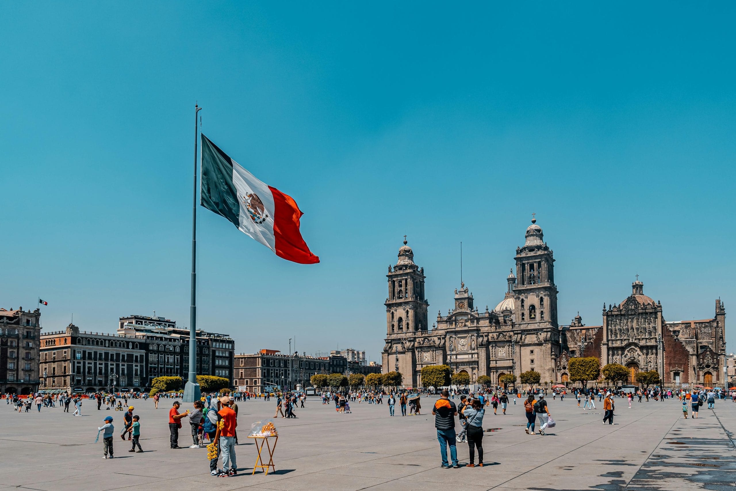 Historic Zócalo plaza in Mexico City with Metropolitan Cathedral and a large Mexican flag flying. Tourists explore the expansive square on a clear blue day. Perfect central location for any Mexico City guide featuring colonial architecture and cultural landmarks in the heart of the capital.